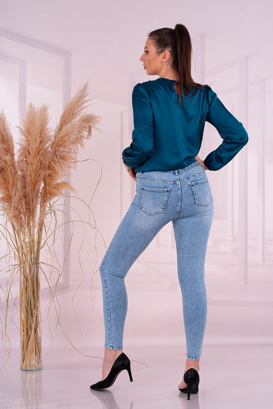 A female model poses with hands on hips looking over her shoulder wearing a teal satin long-sleeve blouse and light blue skinny denim jeans in a pale pink room with tall pampas grass in a glass vase