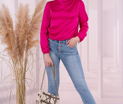 Model standing with one hand in jean pocket against a white room backdrop with pampas grass holding a woven chain-strap bag while wearing a long-sleeve magenta satin draped-neck blouse and light-blue high-waisted skinny denim jeans