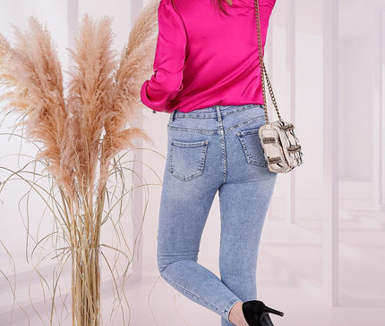 Female model wearing a hot pink satin long sleeve blouse, light blue skinny denim jeans and a cream woven chain strap shoulder bag posed turned away with one leg bent beside a tall glass vase of pampas grass in a white studio