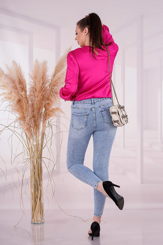 Female model wearing a hot pink satin long sleeve blouse, light blue skinny denim jeans and a cream woven chain strap shoulder bag posed turned away with one leg bent beside a tall glass vase of pampas grass in a white studio