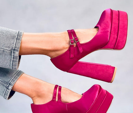 Model wearing fuchsia suede platform Mary Jane heels with double straps and chunky block heels poses with legs raised against a plain grey background