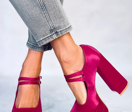 A model wearing grey rolled denim jeans poses with crossed ankles in fuchsia satin platform Mary Jane shoes with block heels against a pale grey floor and backdrop