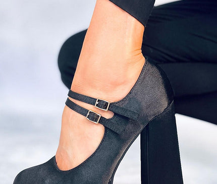 A model wearing black trousers shows a black suede platform Mary Jane pump with two slim ankle straps and a chunky block heel posed against a white floor and wall background