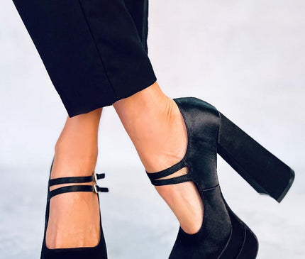 Female model wearing black suede platform Mary Jane block heel shoes with double ankle straps and cropped black trousers standing on a light grey studio floor