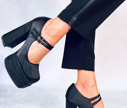 A model in cropped black trousers poses with lifted foot showcasing black satin platform Mary Jane heels with chunky block heels against a light grey studio floor and wall