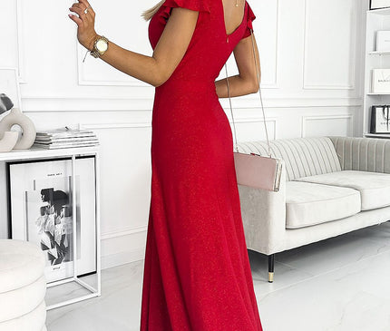 Model wearing a red glitter jersey maxi dress with cap sleeves and flared skirt stands with her back turned in a white panelled room with a grey velvet sofa and a modern white side table with decor