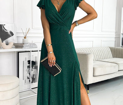 Female model standing in a white paneled room with a cream sofa and framed photos wearing a sparkly forest green wrap-front jersey evening gown with flutter sleeves and high side slit holding a black clutch