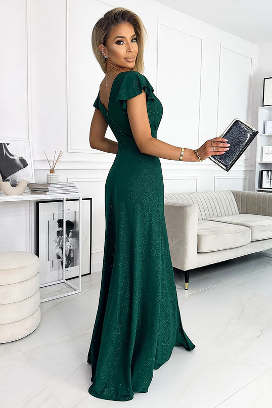Model wearing a green sparkly floor-length mermaid-cut gown with flutter sleeves and a V-back stands sideways looking over her shoulder in a white room with a cream sofa, console table with framed art, books, a reed diffuser and a round ottoman