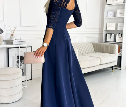 Woman model wearing a navy blue crepe A-line evening dress with lace three-quarter sleeves and a keyhole back turned over her shoulder holding a blush clutch in a white panelled room with a cream sofa and shelves of shopping bags