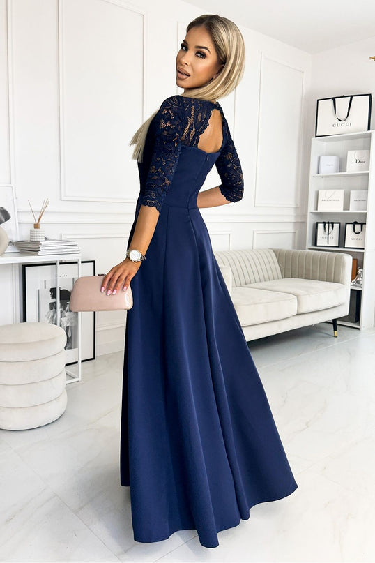Woman model wearing a navy blue crepe A-line evening dress with lace three-quarter sleeves and a keyhole back turned over her shoulder holding a blush clutch in a white panelled room with a cream sofa and shelves of shopping bags