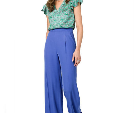 Female model wearing a green floral chiffon V-neck top with ruffle sleeves and cobalt blue high-waisted wide-leg trousers stands against a white background with one hand on her thigh and her head turned to the side