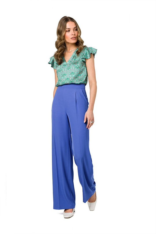 Female model wearing a green floral chiffon V-neck top with ruffle sleeves and cobalt blue high-waisted wide-leg trousers stands against a white background with one hand on her thigh and her head turned to the side