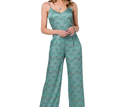 A female model stands front-on against a white background wearing a green patterned satin jumpsuit with spaghetti straps, a V-neckline and wide-leg cut with her hands clasped at her waist