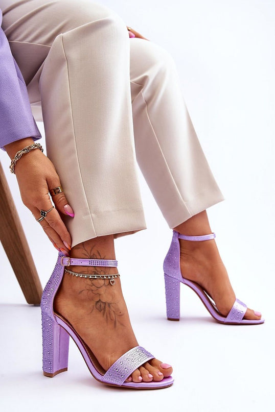 Female model seated on a wooden stool showcases lilac synthetic open-toe block-heel sandals with rhinestone-embellished ankle straps against a white studio backdrop
