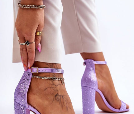 Model adjusts lilac suede open-toe block-heel sandals with rhinestone embellishments against a white studio background