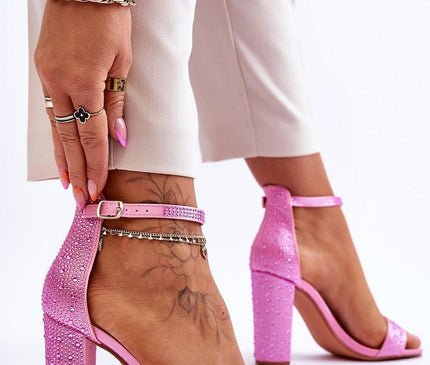 Model wearing pink suede open-toe block heel sandals with ankle strap and rhinestone embellishments adjusting the strap against a plain white background