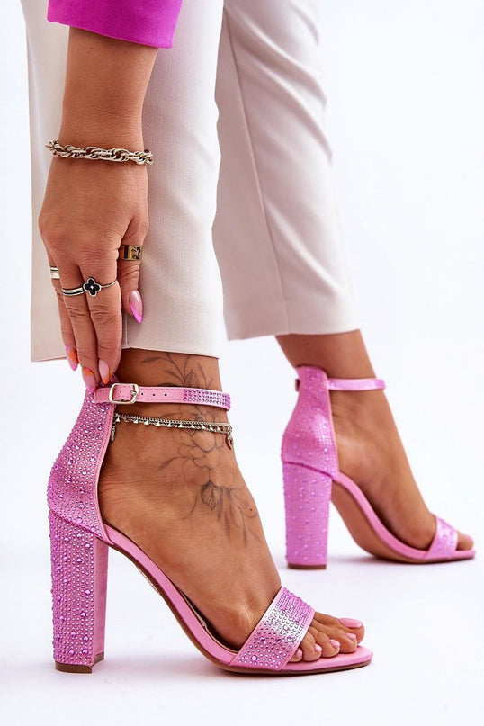 Model wearing pink suede open-toe block heel sandals with ankle strap and rhinestone embellishments adjusting the strap against a plain white background