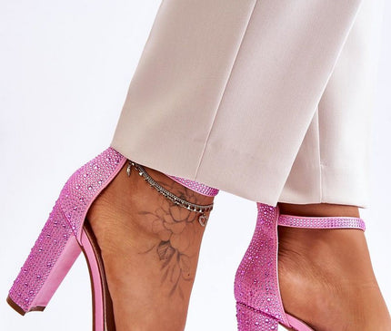 A model wearing beige cropped trousers poses against a white background showcasing pink rhinestone-embellished block-heeled sandals with ankle straps