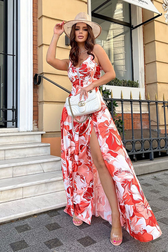 Model wearing a red and white floral satin maxi dress with spaghetti straps and thigh-high slit standing on marble steps outside a beige stone building with black railings and a window while adjusting a straw hat and holding a white shoulder bag
