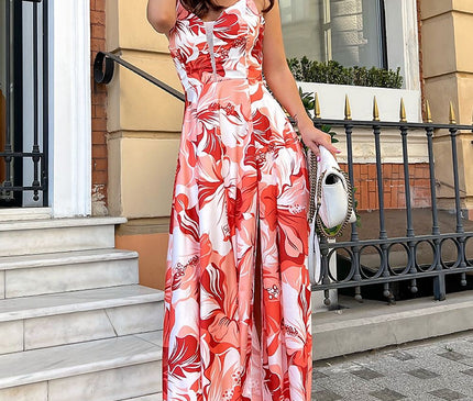 Model in a red and white floral satin spaghetti-strap maxi dress with a front slit holds a straw hat while standing on marble steps beside a wrought iron railing