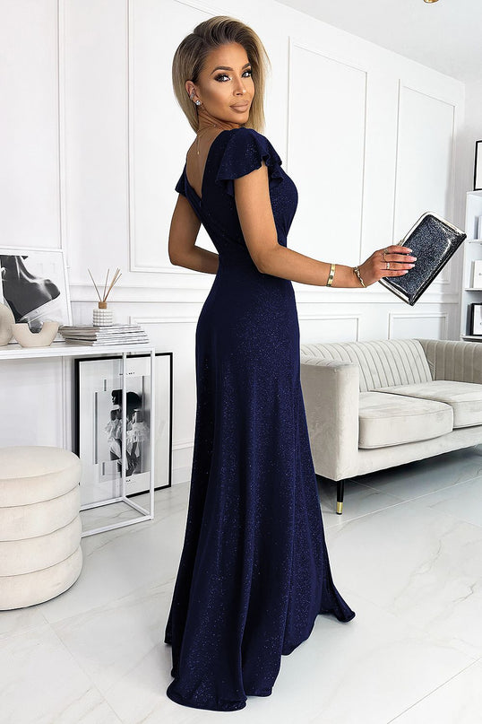 A model holds a black glitter clutch and looks over her shoulder wearing a floor-length navy blue glitter jersey evening dress with V-back and flutter sleeves in a bright room with white paneled walls, cream sofa and framed prints