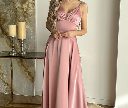 A blonde model stands in a minimalist room with herringbone wood flooring and a marble-top console holding candles wearing a dusty pink satin spaghetti-strap V-neck maxi dress with a fitted bodice and flowing skirt