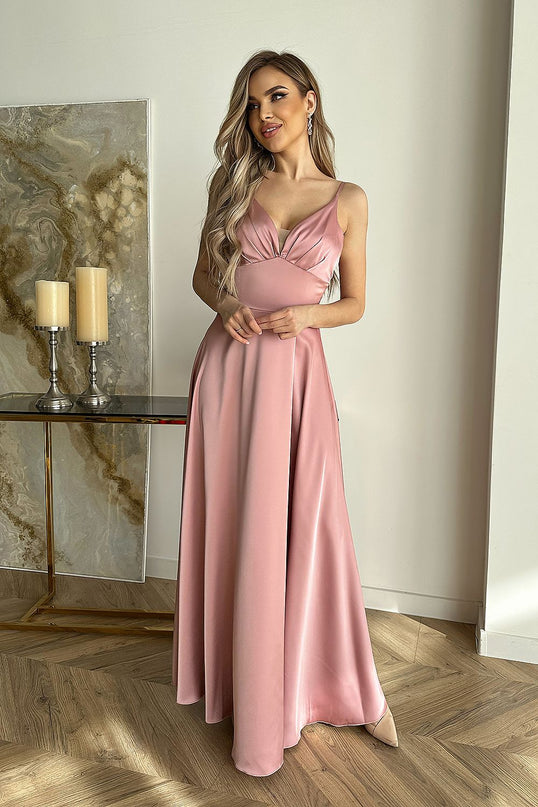 A blonde model stands in a minimalist room with herringbone wood flooring and a marble-top console holding candles wearing a dusty pink satin spaghetti-strap V-neck maxi dress with a fitted bodice and flowing skirt