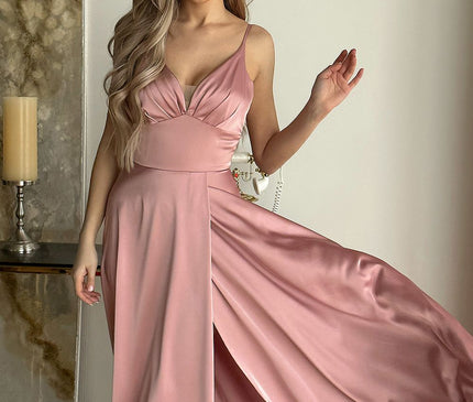 Female model stands holding the flowing skirt of a silky pink satin spaghetti-strap maxi dress with V-neckline against a white wall beside a marble-effect canvas and tall candle on a wooden console