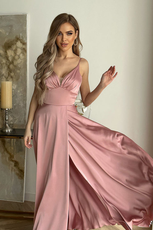 Female model stands holding the flowing skirt of a silky pink satin spaghetti-strap maxi dress with V-neckline against a white wall beside a marble-effect canvas and tall candle on a wooden console