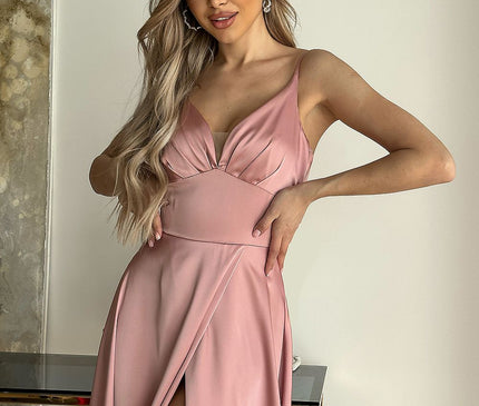 A blonde model stands in front of a cream wall beside a marble-textured painting and a glass-topped gold table wearing a pink satin sleeveless V-neck evening gown with a thigh-high slit and spaghetti straps