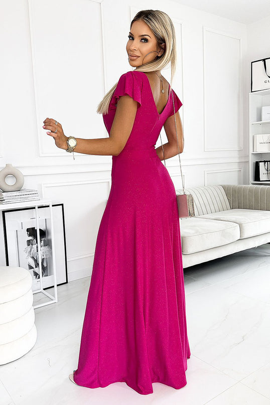 A female model wearing a flowing lightweight fuchsia maxi dress with flutter sleeves and a V-back cut poses turned to the side over her shoulder in a white living room with a sofa framed art and a side table