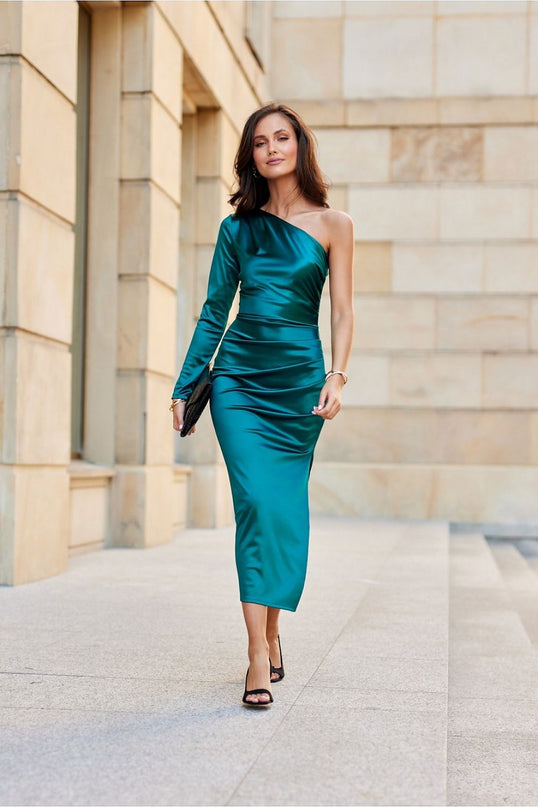 A female model walks forward wearing an emerald green satin one-shoulder fitted midi dress against a beige stone building backdrop