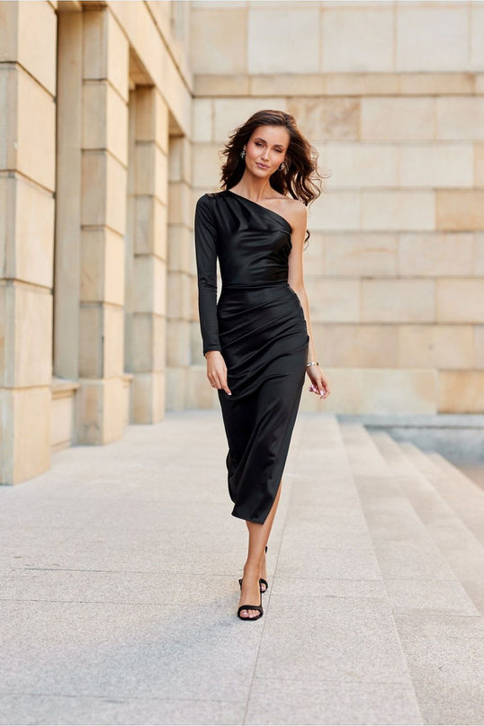 Female model walks in a fitted black satin one-shoulder midi dress with side slit against a backdrop of beige stone columns and steps