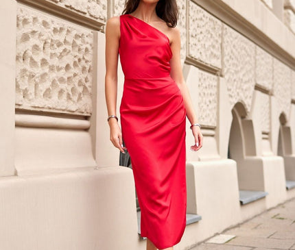 Female model wearing a red satin one-shoulder midi dress with a fitted cut stands poised against a cream textured stone wall on a city pavement
