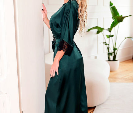 A blonde model in a side pose wears a long emerald green satin robe with black lace trim and a tie belt barefoot while leaning against a white paneled wall in a bright room with wooden flooring and a potted plant