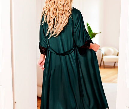 A blonde model stands barefoot in a bright room wearing a floor-length emerald green satin wrap robe with black lace trim at cuffs and hem holding the flowing skirt over a wooden floor with a cream armchair and potted plant in the background